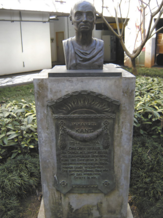 Imagem de busto sobre uma base retangular. A parte frontal da base contém uma placa com inscrições. O busto representa um homem idoso, com expressão séria.