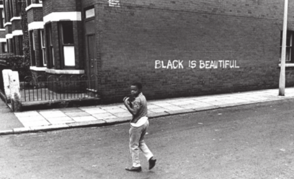 Imagem em preto e branco de uma criança negra atravessando uma rua. Na parede lateral de um sobrado, há uma frase em inglês.