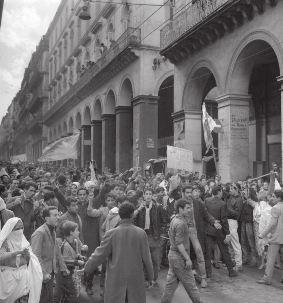 Imagem em preto e branco de manifestantes com faixas e cartazes. Eles estão em frente a um edifício com quatro pavimentos e galerias.