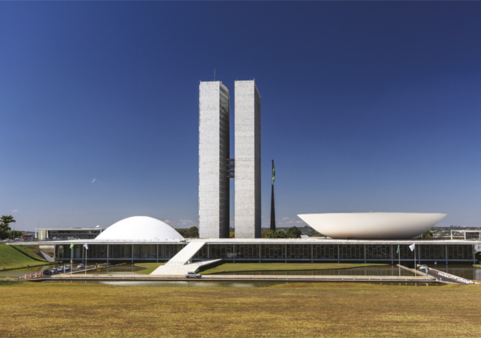 Imagem de edifício com paredes de vidro. Duas rampas levam à entrada e ao terraço superior, onde há duas torres entre um elemento côncavo, à direita, e um elemento convexo, à esquerda. Em frente ao edifício, há um espelho d'água.