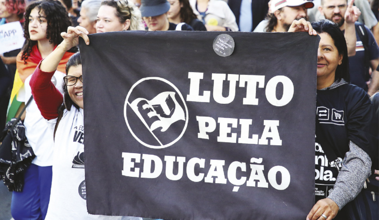 Imagem de manifestantes. Duas mulheres seguram uma faixa preta com a seguinte frase: Eu luto pela educação.