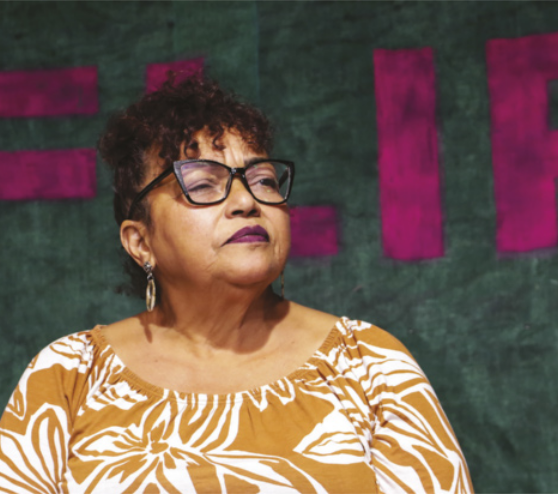 Fotografia de Débora Silva. Ela é negra, possui cabelos cacheados e usa óculos, brincos e blusa com estampa floral.