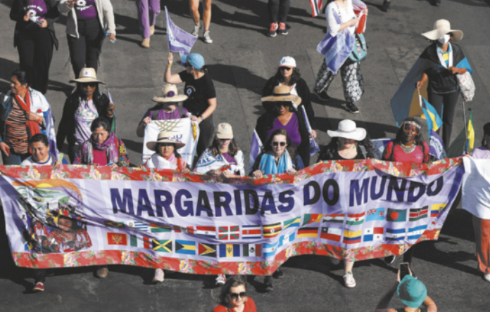 Imagem de mulheres em um protesto. Elas caminham carregando uma faixa com diversas bandeiras e o título Margaridas do Mundo.
