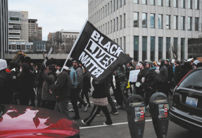 Imagem de manifestantes caminhando em uma rua com diversos edifícios.