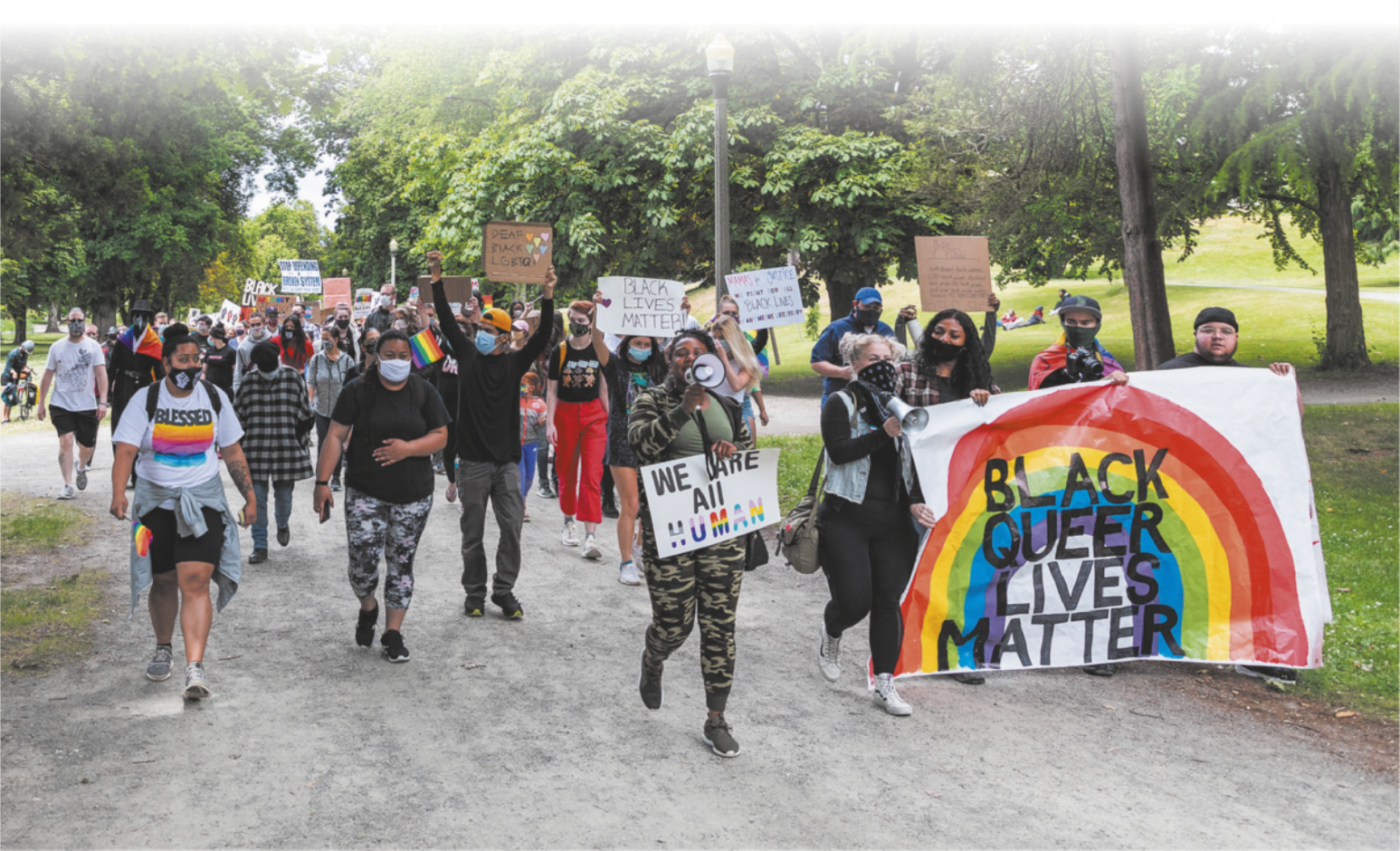 Imagem de manifestantes em um parque. Eles carregam diversos cartazes, um deles contém a figura de um arco-íris.