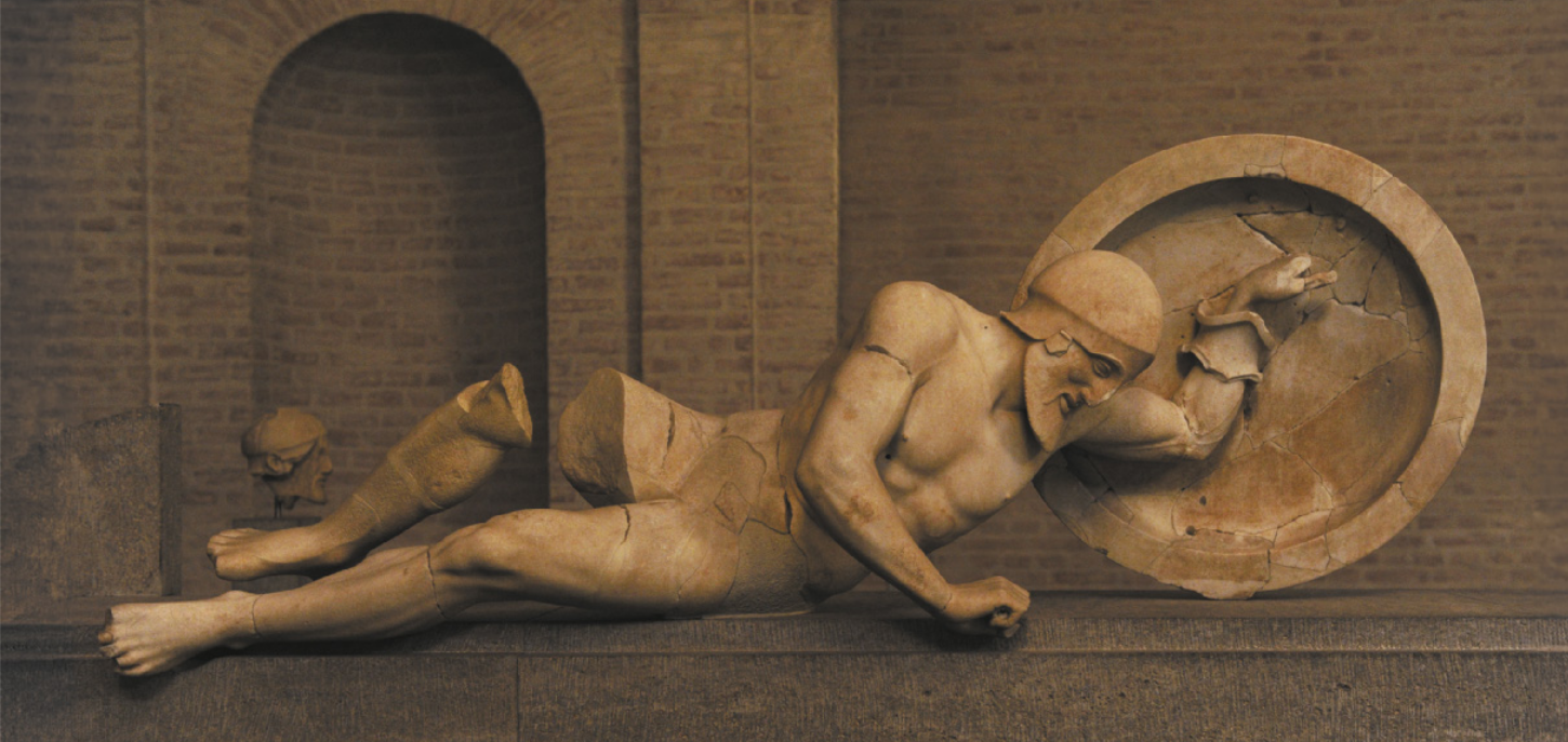 Imagem de escultura que representa um guerreiro derrotado. Ele está deitado, com o tronco voltado para baixo, a mão direita apoiada no chão e o braço esquerdo encaixado na alça do escudo. A região da coxa e do joelho direito está fragmentada.