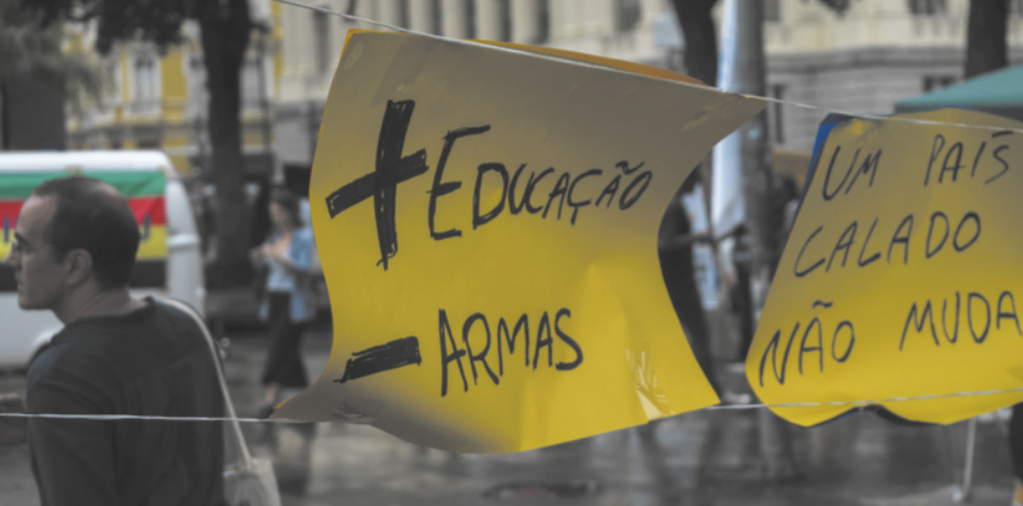Imagem de cartazes pendurados em uma praça. Eles contêm as seguintes frases: Mais educação, menos armas. Um país calado não muda.