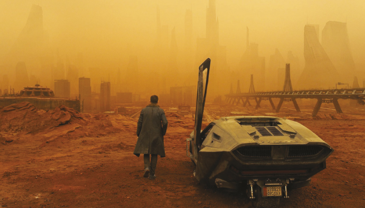 Imagem da cena de um filme que mostra um homem ao lado de um carro. Ele caminha em direção ao uma paisagem desértica e enevoada, com diversos prédios.