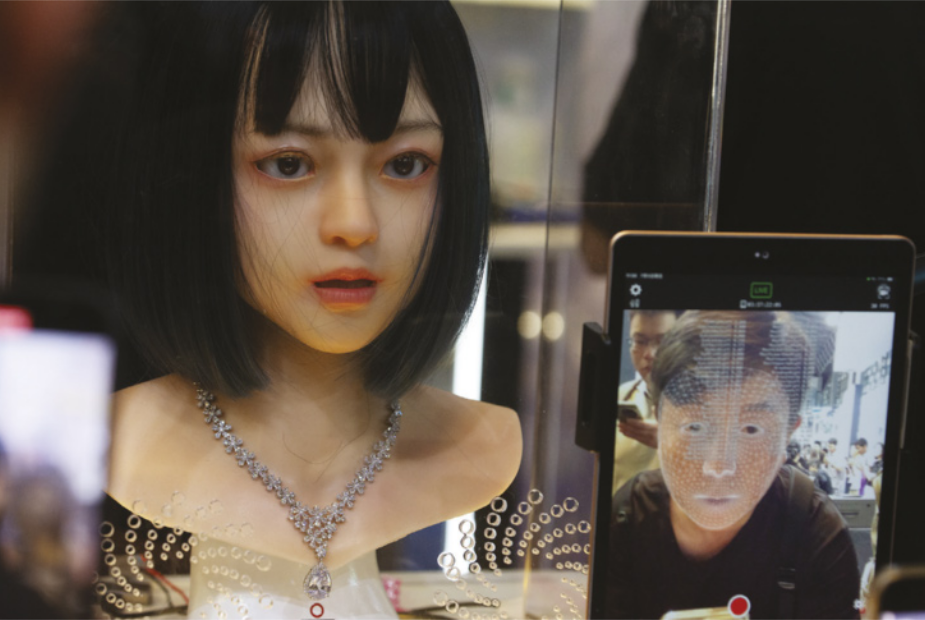 Imagem de máscara feminina em um expositor. Ela possui cabelo liso com franja, está com a boca entreaberta e usa um colar. Na parte frontal do expositor, um tablet mostra a imagem de uma pessoa.