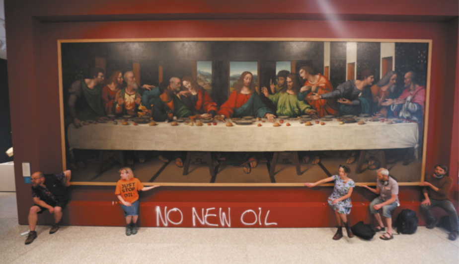 Imagem de ativistas em um museu. Eles estão sentados em um banco e tocam a moldura de um quadro. A parte frontal do banco contém uma pichação em inglês.