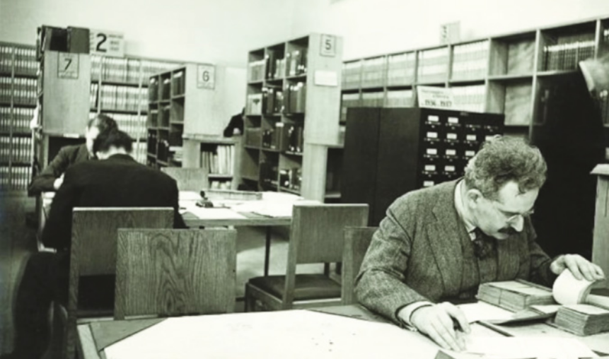 Fotografia em preto e branco de Walter Benjamin. Ele está sentado diante de uma mesa, lendo livros e documentos. Atrás dele, dois homens estão lendo. Ao redor deles, há diversas estantes com livros.