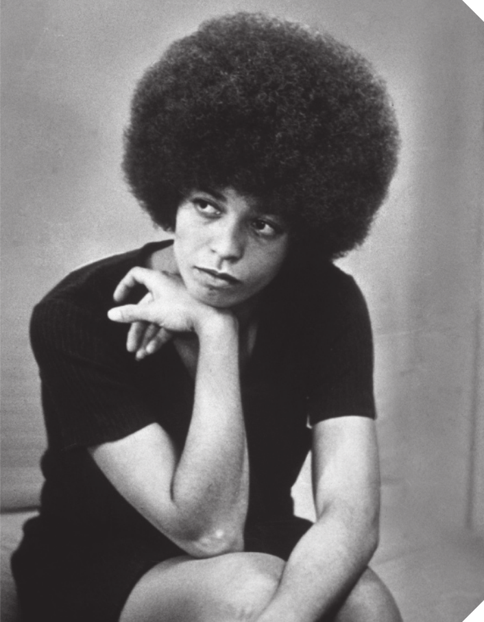 Fotografia em preto e branco de Angela Davis. Ela é negra, possui cabelo crespo armado, usa vestido curto e está sentada, com o queixo apoiado sobre o dorso da mão direita.