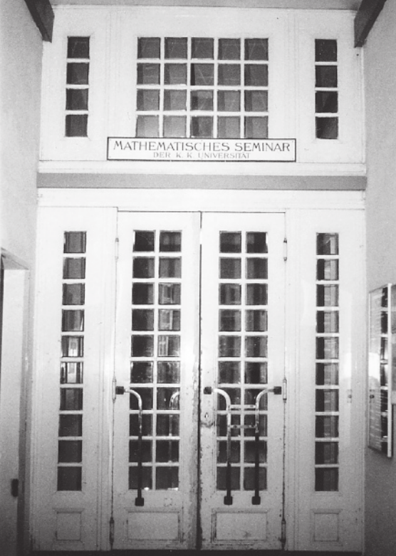 Fotografia em preto e branco de porta dupla, com vidros quadrados e puxadores. Acima da porta, há uma placa.