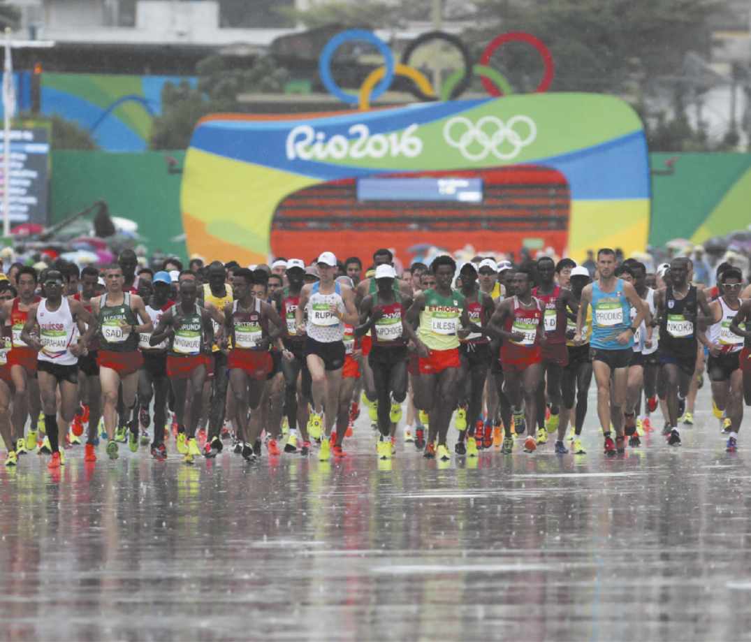 Imagem de maratonistas correndo na chuva. Eles usam tênis, shorts, regata e placa de identificação no peito.