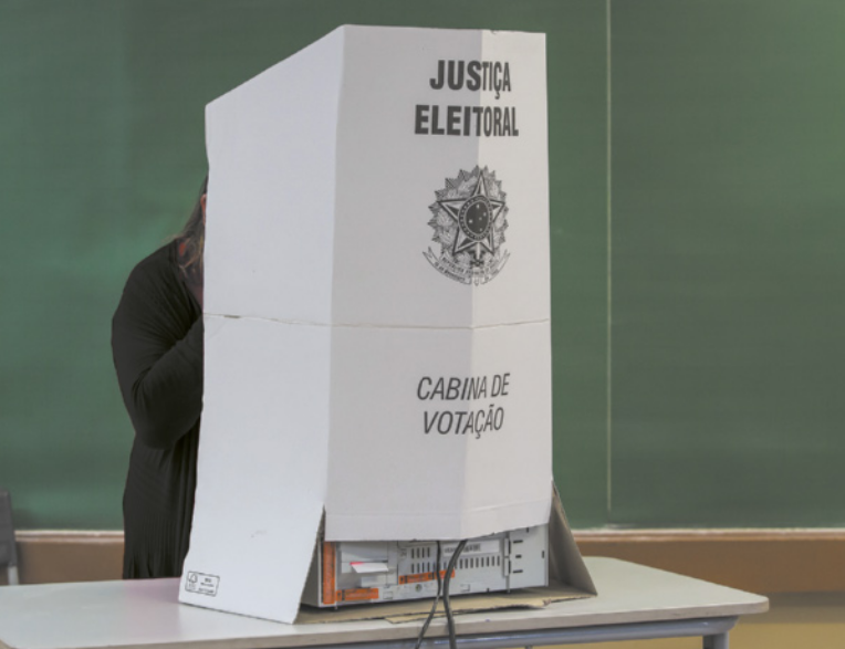 Imagem de mulher votando por meio da urna eletrônica. A urna está em uma cabine de votação, montada sobre uma mesa.