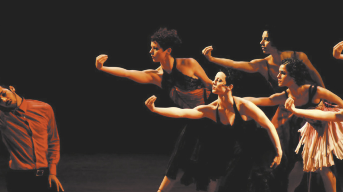 Imagem de uma cena de espetáculo de dança. Um grupo de bailarinas, à direita, está com o tronco curvado, o braço estendido para frente e a mão curvada em forma de concha, com o dorso para fora. Diante delas, um bailarino pende a cabeça para o lado direito.