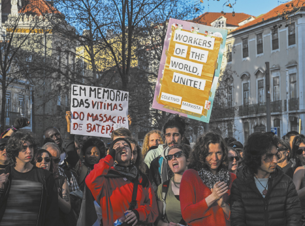 Imagem de manifestantes em uma área cercada por edifícios. Eles carregam dois cartazes, um deles com a frase em inglês: 'Workers of the world, unite!', coletivo marxista. O outro cartaz contém a seguinte frase: 'Em memória das vítimas do massacre de Batepá'.