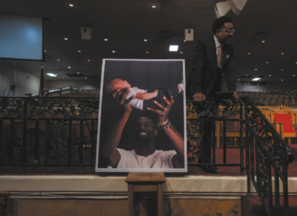 Imagem de cartaz exposto em um auditório. O cartaz mostra um homem negro sorrindo e erguendo um bebê deitado.