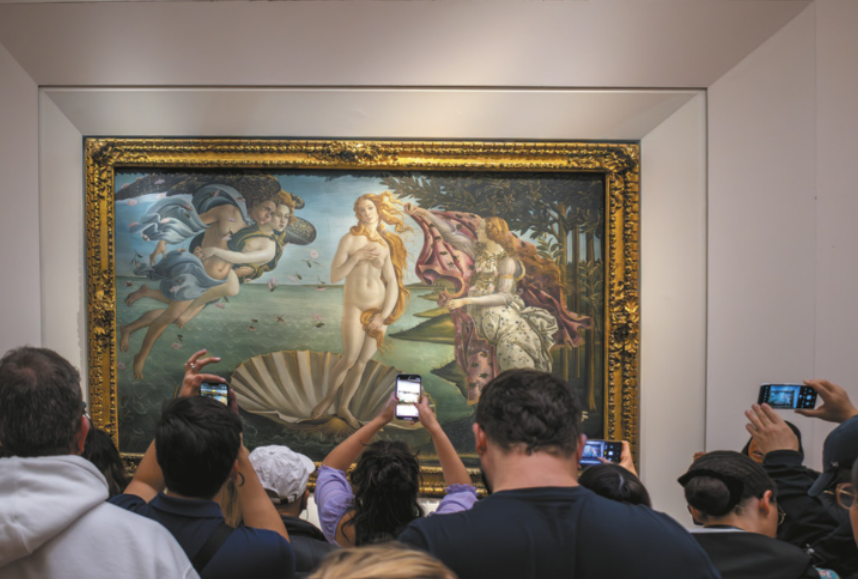 Imagem de pessoas utilizando smartphones para fotografar um quadro, que retrata Vênus nua. Ela está em pé sobre uma concha que flutua no mar. À esquerda, dois serem alados estão abraçados e sopram em direção à concha. À direita, uma deusa estende um manto floral para Vênus.