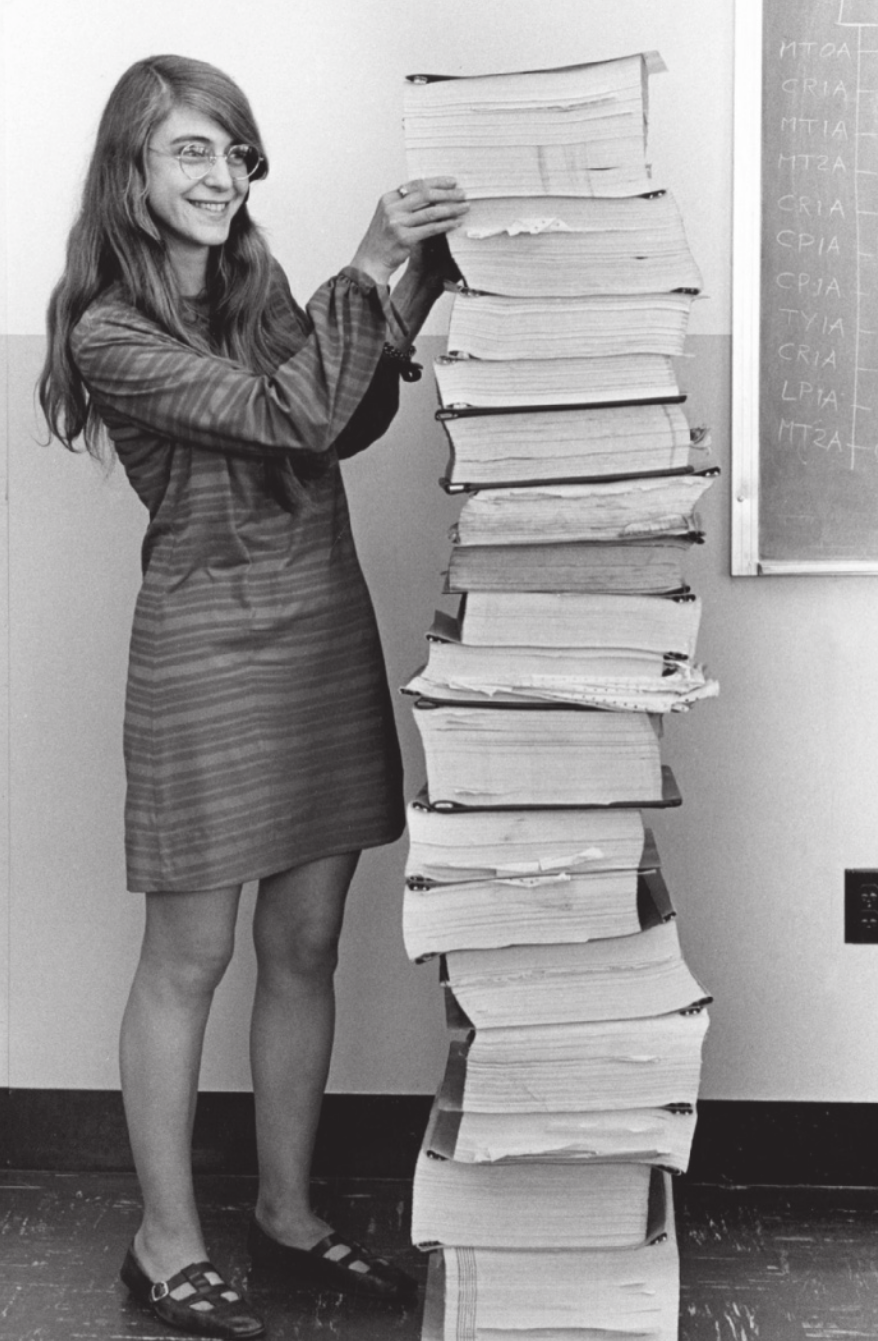 Fotografia em preto e branco de Margaret Hamilton. Ela é branca, possui cabelos longos, usa óculos e vestido, está sorrindo e organiza uma pilha de livros do tamanho dela.