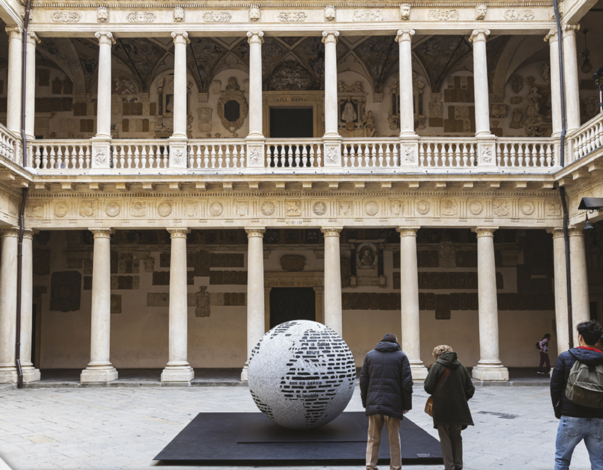 Imagem do pátio interno de uma universidade. O edifício possui dois pavimentos, cada um com galerias cercadas por colunas. No centro do pátio, pessoas estão ao redor de um globo terrestre.