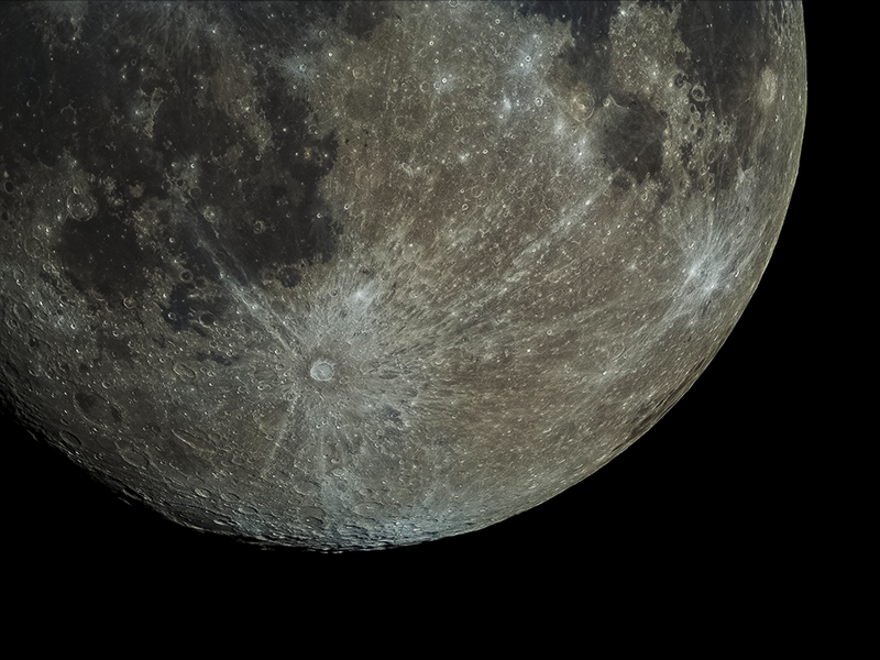 Fotografia de parte da Lua com destaque para uma cratera na parte inferior. Ela tem o formato circular, com coloração esbranquiçada e brilhante. Dela, saem vários raios, também brilhantes, que se estendem por várias partes da superfície lunar.