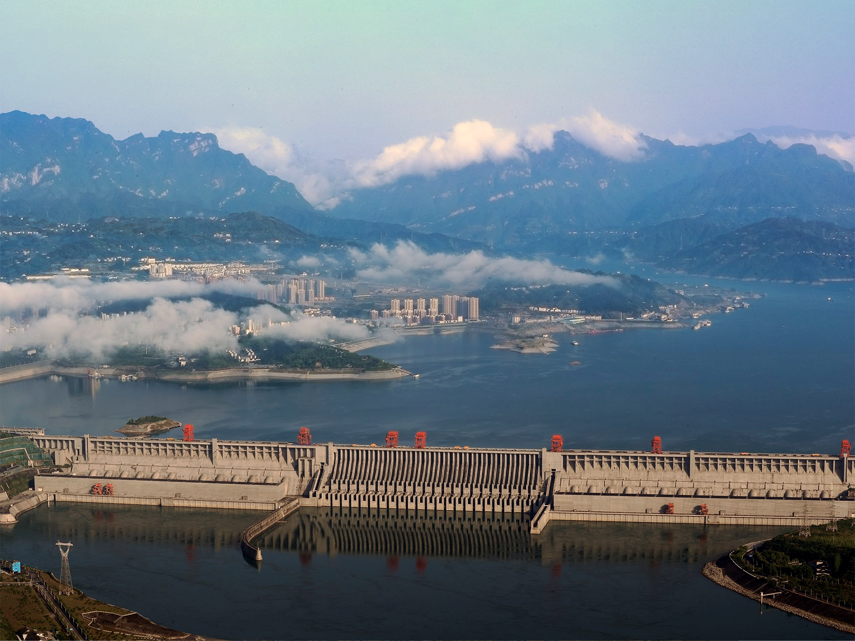Fotografia de uma grande barreira de concreto dividindo as águas de um rio, correspondente a uma usina hidrelétrica. Na base da barreira, há aberturas para a passagem da água. Ao fundo, há vários prédios e névoa encobrindo parte da paisagem. Mais ao fundo, há áreas montanhosas cobertas por vegetação e grandes nuvens acima.