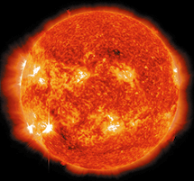 Fotografia de satélite do Sol. Ele tem formato esférico com feixes amarelados em sua borda. A superfície tem coloração alaranjada, com tons de vermelho e manchas amarelas brilhantes, de onde saem feixes luminosos.