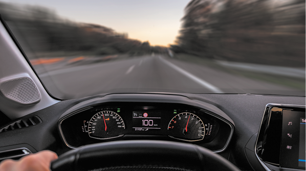 Fotografia do painel de um carro visto a partir do volante. No painel há dois ponteiros indicadores, um deles o velocímetro e no meio está o número 100. À frente, uma estrada é vista a partir do para-brisa.