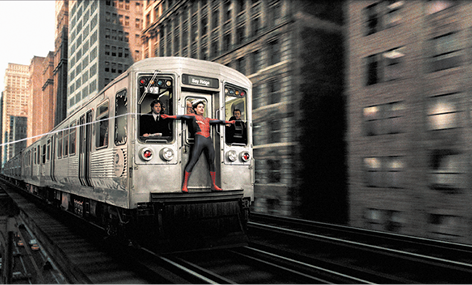 Fotografia de uma cena de um filme, com um homem usando roupa do Homem-Aranha, azul e vermelho. Ele está tentando parar um trem em movimento, está em pé sobre uma base da parte frontal do trem, com os braços estendidos segurando dois cabos de teias. Ao lado do trilho do trem há prédios que estão desfocados.