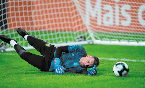 Fotografia de um goleiro em posição de queda no gramado. Ele está deitado de lado com as pernas para cima, um dos braços está estendido e há uma bola logo ao lado.