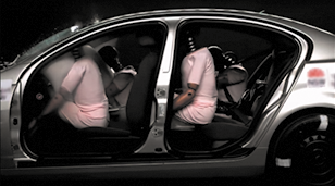 Fotografia de uma simulação de colisão de um carro com ativação do airbag. O carro é visto a partir da lateral, sem as portas e possui 4 bonecos que simulam pessoas. No banco do motorista e do passageiro da frente os airbags estão acionados e os bonecos estão inclinados sobre eles. No banco traseiro, um dos bonecos está sentado no banco com o cinto de segurança e o outro boneco está inclinado sobre o banco da frente.