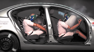 Fotografia de uma simulação de colisão de um carro com ativação do airbag. O carro é visto a partir da lateral, sem as portas e possui bonecos sentados, que simulam pessoas. Na frente, no banco do passageiro, o airbag está ativado, e o boneco está inclinado. No banco traseiro, os bonecos estão sentados, e estão projetados com o pescoço e braços para a frente do encosto do banco. Há fumaça em torno do carro.