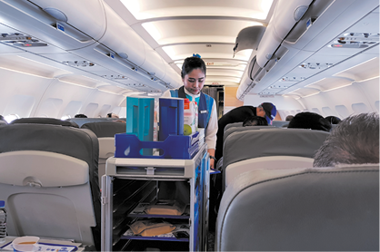 Fotografia de uma aeromoça empurrando um carrinho com garrafas de bebidas e pratos de alimentos, em um corredor de um avião.