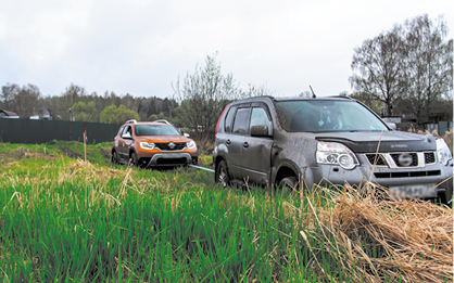 Fotografia de uma estrada de terra com dois carros. O carro da frente é cinza e está puxando o carro de trás, que é laranja, com o auxílio de uma corda que está presa nos dois carros. Em torno da estrada há grama e algumas árvores ao fundo.