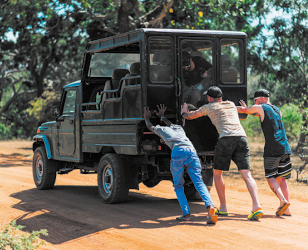 Fotografia de três homens empurrando a traseira de um veículo jipe em uma estrada de terra rodeada por vegetação.