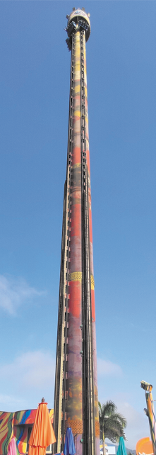 Fotografia de um brinquedo de parque de diversões formado por uma torre colorida e alta com bancos nos quatro lados da torre. Os bancos estão sobre os trilhos verticais da torre e está posicionado na parte mais alta.