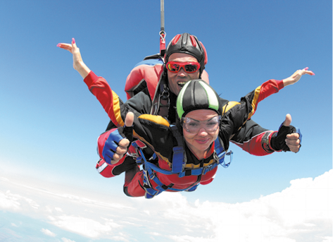 Fotografia de duas pessoas em um salto de paraquedas. Uma delas está por cima, enquanto a outra está por baixo em um mesmo paraquedas. Ambas estão sorrindo, usam capacete e óculos e estão com os braços abertos. Ao fundo está o céu azul com nuvens.