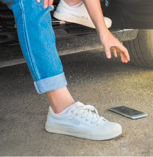 Fotografia de uma pessoa sentada no banco de um carro, com a porta aberta. Ela é vista dos joelhos para baixo e um dos pés está apoiado na rua, enquanto o um braço está estendido na direção de um celular que está caído no chão.