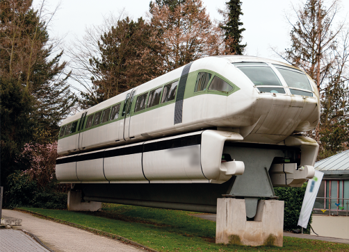 Fotografia de parte de um trem nos trilhos. Ele é branco e nas laterais há janelas e portas. O trem e os trilhos estão expostos sobre duas bases de concreto em um gramado com árvores ao fundo.