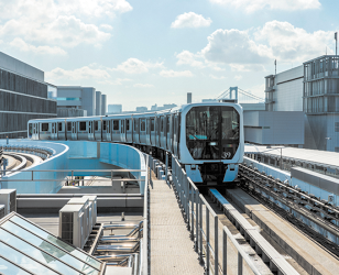 Fotografia de um trem sobre os trilhos durante uma curva. Ele é azul e ao redor há estruturas metálicas próximas dos trilhos e edifícios ao fundo.