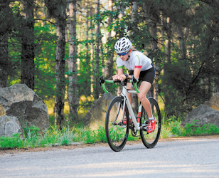 Fotografia de uma ciclista pedalando em uma estrada ao ar livre, cercada por pedras e vegetação. Ela usa capacete, óculos, camiseta, shorts e sapatilha de ciclismo.