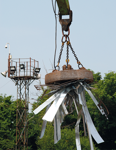 Fotografia de um guindaste no alto, elevando várias peças metálicas que estão grudadas na extremidade do guindaste, a qual possui formato circular. Ao fundo há uma torre e várias árvores.