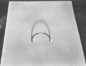 Fotografia de uma superfície contendo um fio que sai de um determinado ponto e volta para a superfície em outro ponto formando um arco. Há partículas metálicas na superfície compondo um desenho circular em volta do fio.