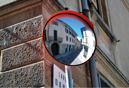 Fotografia de um espelho curvo anexado no vértice da fachada de um edifício. O espelho reflete o cruzamento entre duas ruas cercadas por edifícios.