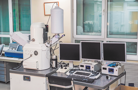 Fotografia de uma grande máquina apoiada em uma mesa metálica, ao lado de um computador com 3 telas. A máquina tem uma base retangular com um tubo cilíndrico em cima, e nas laterais há outras hastes e mais tubos.