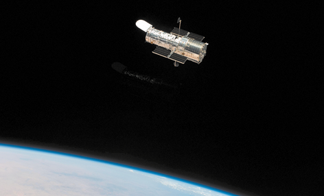 Fotografia de um telescópio visto no espaço. Ele tem o formato cilíndrico com placas nas laterais. Abaixo está parte de um planeta com coloração azul.