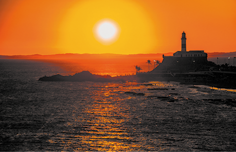 Fotografia do sol se pondo no horizonte. Em primeiro plano está o mar com uma parte de terra mais ao fundo, onde há um farol acima de uma montanha. O céu tem cor alaranjada com o sol amarelo brilhante em destaque no centro.