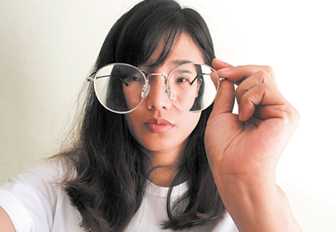 Fotografia do busto de uma mulher, segurando um óculos em frente ao rosto, a uma certa distância. Nas lentes do óculos o rosto é visto em um tamanho menor.