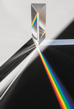 Fotografia de um prisma de base triangular feito de vidro transparente. Há uma luz branca que incide sobre o prisma, que a dispersa em um feixe de luz colorida, com tons de vermelho, amarelo, verde e tons de azul.